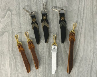 May include: A collection of leather keychains in various colors, including black, brown, and white. Each keychain features a metal clasp and decorative braided or stitched leather details. The keychains are arranged on a light gray wooden surface.