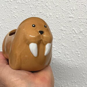 May include: A glossy, light brown ceramic planter shaped like a walrus. The walrus features two prominent white tusks, a small black nose, and two dark eyes. The top of the walrus head has an open cavity for holding small items or plants.