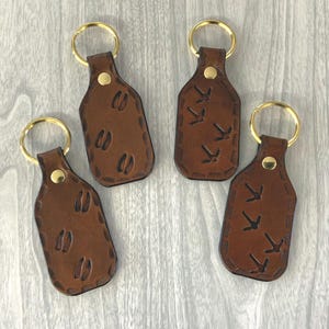 May include: Four brown leather keychains with gold-colored key rings. Two keychains feature deer track designs, and the other two have bird track designs. Each keychain has a gold-colored rivet.