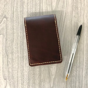 May include: A brown leather notebook with a yellow stitch detail. The notebook is lying on a light wood surface next to a black and clear pen.