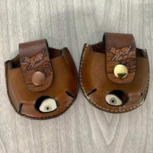 May include: Two round brown leather pouches featuring a stamped deer design on the front flap. Each pouch has a circular cutout at the bottom and a metallic snap closure. The left pouch has a copper-toned snap, while the right has a gold-toned snap.