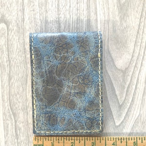 May include: A blue and gray leather wallet with a stitched edge. The wallet has a unique pattern of light blue lines on a dark gray background.