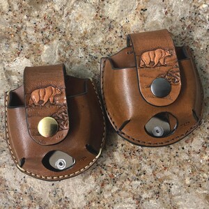 May include: Two brown leather pouches with a bear carving on the flap. Each pouch has a snap closure and a circular opening at the bottom. The pouches are on a speckled brown surface.