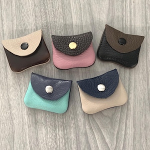 May include: A collection of five small, leather coin purses in various colors. Each purse has a flap closure with a button. Colors include brown, pink, black, teal, and beige, with contrasting flap colors. The purses are arranged on a light wood-grain surface.