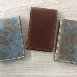 May include: Three leather wallets, one brown and two with a distressed blue and gray pattern. The wallets have a light brown stitching around the edges.