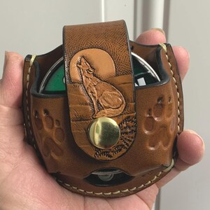 May include: A brown leather coin purse with a detailed embossed design of a howling wolf under a full moon. The purse has a snap closure and paw print accents. The purse is designed to hold coins.