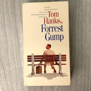 May include: A VHS tape for the movie "Tom Hanks is Forrest Gump." The cover features the title in red and blue, with a man sitting on a bench. The text "The world will never be the same" is also visible.