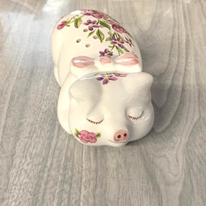 May include: A white ceramic piggy bank with a pink bow and purple flowers painted on it. The piggy bank is lying on its side with its eyes closed.