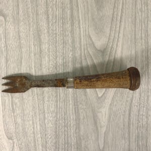 May include: An antique, rusty metal fork with a wooden handle. The fork has three tines and a worn, aged appearance. The handle is cylindrical and appears to be made of wood with a metal cap. The tool is likely used for kitchen or dining purposes.