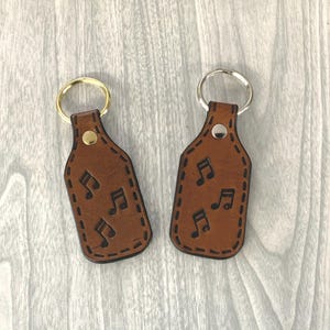 May include: Two brown leather keychains with silver and gold key rings. Each keychain has a stitched border and features a design of musical notes.