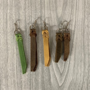May include: Five leather keychains with silver clasps. The keychains are braided and come in green, brown, tan, dark green, and brown.