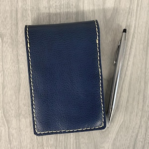 May include: A navy blue leather notebook with white stitching and a silver pen. The notebook is rectangular and closed, with a textured surface. The pen is silver and sleek, positioned next to the notebook.