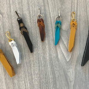 May include: A collection of seven leather zipper pulls in various colors including mustard yellow, white, black, brown, turquoise, and dark gray. Each pull features a braided leather knot attached to a metal lobster clasp or a metal hook.