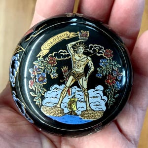 May include: A black, round decorative box with a detailed illustration of the Colossus of Rhodes. The image features a figure holding a crown, surrounded by floral designs in red, blue, and gold. The word "COLOSSUS" is written on a banner.