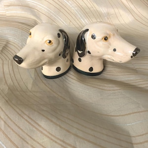 May include: A pair of ceramic salt and pepper shakers shaped like Dalmatian dog heads. The shakers are off-white with black spots, black ears, and black noses. The eyes are yellow. The bases are black.