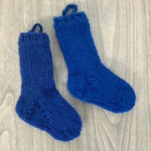 May include: Two knitted blue socks with a loop at the top for hanging. The socks are made of a chunky yarn and have a ribbed cuff. The socks are displayed on a light gray wooden surface.