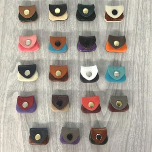 May include: A collection of small, colorful leather pouches with snap closures. The pouches are in various colors, including brown, black, pink, blue, and orange. Each pouch has a contrasting flap and a metal snap. The pouches are arranged on a light wood surface.