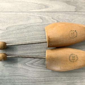 May include: A pair of wooden shoe stretchers with a spring mechanism. The stretchers are connected by a metal spring and have a round wooden knob on each end. The stretchers have the text "SALA" and "MANAR" printed on them.