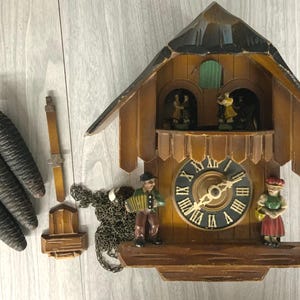 May include: A wooden cuckoo clock featuring a dark brown gabled roof, a black clock face with gold Roman numerals, and two figurines on the front. Included are three dark pinecone-shaped weights, a metal chain, and two small wooden components.