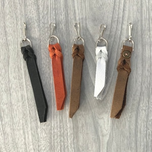 May include: Five leather keychains with different colored braided leather straps and silver key rings. The keychains are brown, black, white, orange, and a darker brown.