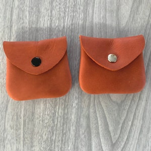 May include: Two small, brown leather coin purses with flap closures and metal snaps. The purse on the left has a black snap, and the purse on the right has a silver snap.