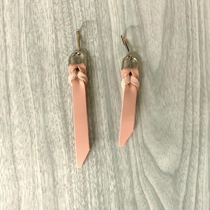 May include: A pair of blush pink leather earrings. Each earring features a braided top connected to a gold-colored hook. The leather strips are rectangular with a diagonal cut at the bottom. The earrings are displayed on a light gray wooden surface.
