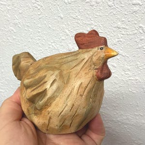 May include: A carved wooden hen figurine with a detailed, textured surface. The hen is primarily tan with a red comb and wattle, and a yellow beak. The figurine is held in a hand against a white textured background.