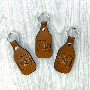 May include: Three brown leather keychains with silver key rings. Each keychain features a raised design of two rams' heads.