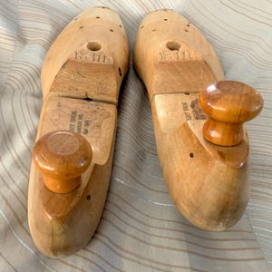 May include: Two wooden shoe forms with a light brown finish. Each form has a rounded knob at the toe and a rectangular piece of wood attached to the heel. The forms are designed to maintain the shape of shoes.