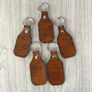 May include: Five brown leather keychains with silver key rings. Each keychain features a paw print design. The keychains are arranged on a light gray wooden surface. The leather has a natural, textured appearance.