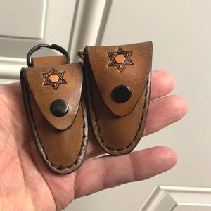 May include: Two brown leather keychains with a Star of David design. Each keychain has a black snap closure and a stitched border. One keychain has a black metal ring.