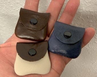 May include: Three small square pouches with rounded bottom corners and fold-over flaps secured by black circular snap buttons. One pouch is dark brown, one is navy blue, and one features a cream-colored body with a dark brown flap.