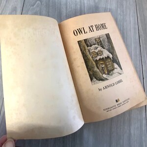 K&ouml;nnte beinhalten: Ein offenes Buch mit dem Titel "OWL AT HOME" von Arnold Lobel. Die Illustration auf dem Einband zeigt ein schneebedecktes Haus in einem Wald. Das Buch stammt von Scholastic Book Services.