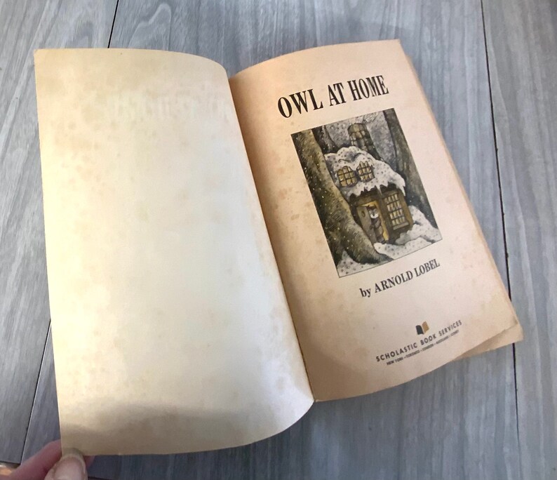 K&ouml;nnte beinhalten: Ein offenes Buch mit dem Titel "OWL AT HOME" von Arnold Lobel. Die Illustration auf dem Einband zeigt ein schneebedecktes Haus in einem Wald. Das Buch stammt von Scholastic Book Services.