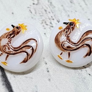 May include: Pair of white, round earrings featuring a brown rooster design with gold accents. The rooster is depicted with a stylized body and a golden comb. These earrings are a unique accessory.
