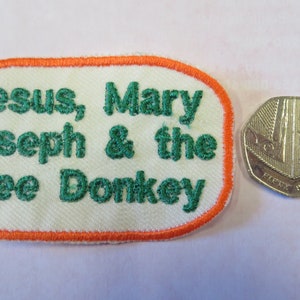 May include: A white fabric patch with an orange border and green embroidered text that reads "Jesus, Mary Joseph & the Wee Donkey".