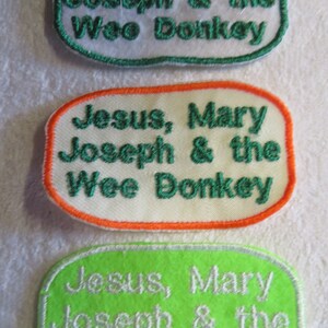May include: Three embroidered patches with the text "Jesus, Mary, Joseph & the Wee Donkey." The patches are oval-shaped with different colour combinations: green border on white, orange border on white, and silver border on green.