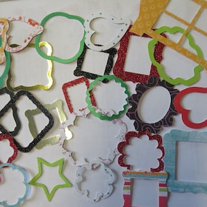 May include: A collection of colourful paper frames in various shapes and sizes, including hearts, stars, and rectangles. The frames are decorated with glitter, patterns, and stripes.