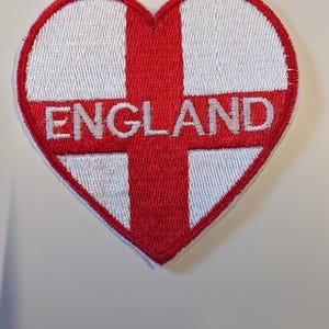 May include: A heart-shaped embroidered patch featuring the white and red cross of the England flag. The word ENGLAND is stitched in white capital letters across the centre of the red cross. The patch has a thick red stitched border.