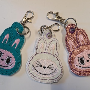 May include: Three embroidered bunny keyrings in teal, white, and glittery rose gold. Each keyring features a bunny face with a mischievous expression, stitched details, and a silver-tone clasp for attaching to keys or bags.