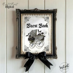May include: A framed print with the words "Guest Book" in gothic script. The artwork features a vintage baby carriage with black flowers and bats. The frame is ornate black, and a black bow is attached below.