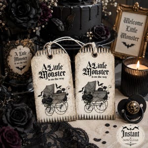 May include: Two rectangular paper tags with a vintage beige finish and black striped edges. Each tag features the text A Little Monster is on the way above a graphic of a baby carriage adorned with flowers and bats, suspended by twine.