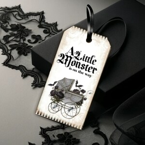 May include: A rectangular paper gift tag with a black ribbon loop, featuring the text A Little Monster is on the way in black gothic font. The design includes a vintage-style baby carriage illustration with dark floral accents and two bats.