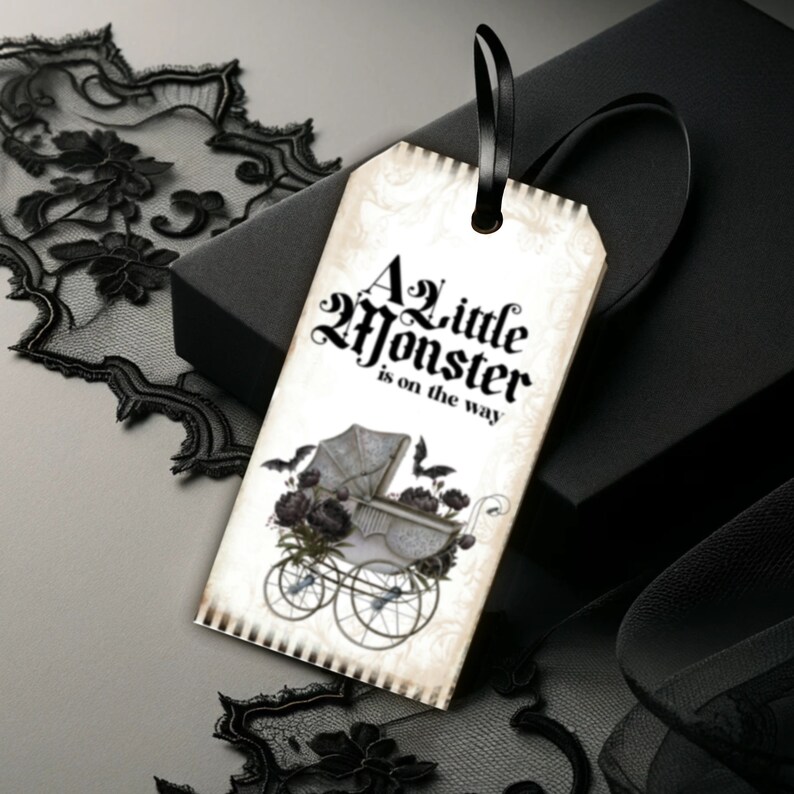 May include: A rectangular paper gift tag with a black ribbon loop, featuring the text A Little Monster is on the way in black gothic font. The design includes a vintage-style baby carriage illustration with dark floral accents and two bats.