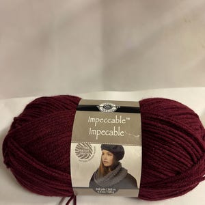 May include: A large ball of burgundy Impeccable yarn with a label featuring a woman wearing a beret and scarf. The label indicates 268 yards and 4.5 ounces of yarn. The yarn is a deep, rich shade of red.