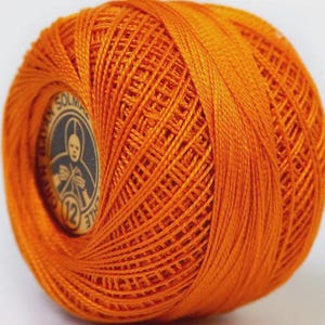 May include: A vibrant orange ball of thread, tightly wound and ready for crafting. The thread is a rich, saturated hue, with a visible woven texture. A circular label with a vintage design is partially visible, adding a touch of classic charm.