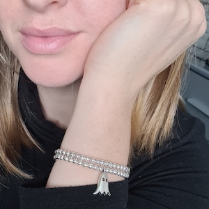 May include: A silver beaded bracelet with a ghost charm. The bracelet is made of small, round silver beads and features a dangling ghost charm with two small eye holes. The bracelet is worn on a wrist.