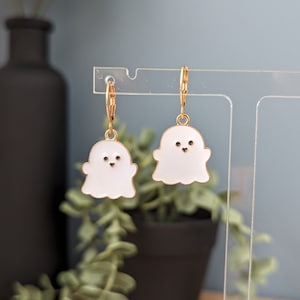 May include: A pair of gold-tone ghost-shaped earrings with white enamel. The earrings have small black eyes and a smiling mouth.