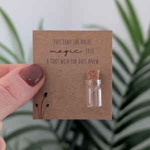 May include: A miniature glass jar with a cork stopper is attached to a brown card. The card features the text "THIS TINY JAR HOLDS MAGIC TRUE A TINY WISH FOR DAYS ANEW". A small black floral design is in the lower left corner.