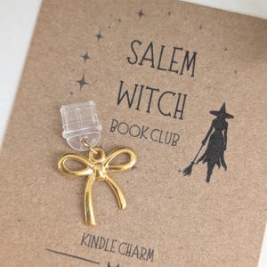 May include: A gold-coloured metal bow charm attached to a clear plastic clip. The charm is displayed on a brown card printed with the text SALEM WITCH BOOK CLUB, KINDLE CHARM, and DAISY MOON GIFTS, along with a black silhouette of a witch.